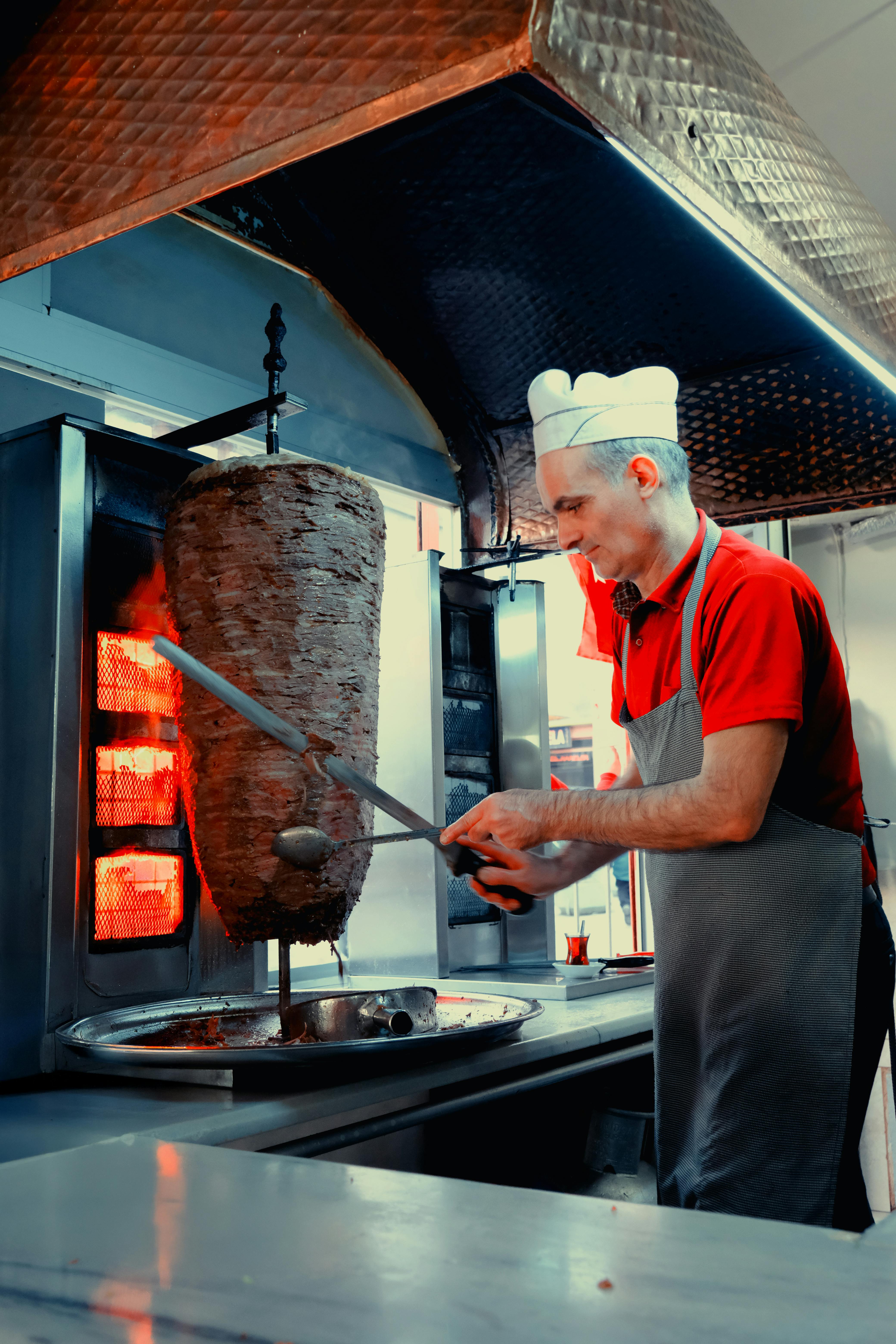 A chef skillfully slices shawarma meat from a large rotisserie skewer in a restaurant kitchen setting.